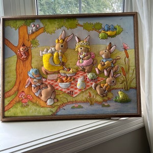 May include: A framed felt appliqu&eacute; picture of a group of bunnies having a picnic in a forest setting. The bunnies are wearing clothes and holding food and musical instruments. There is a frog on a lily pad in a pond and a bird in a nest in a tree.