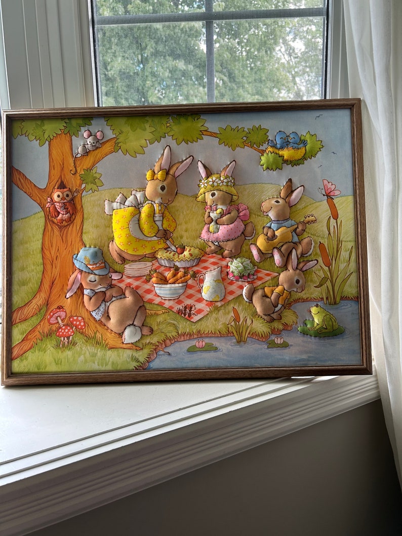 May include: A framed felt appliqu&eacute; picture of a group of bunnies having a picnic in a forest setting. The bunnies are wearing clothes and holding food and musical instruments. There is a frog on a lily pad in a pond and a bird in a nest in a tree.