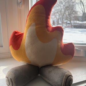 May include: A plush toy in the shape of a campfire flame, with a colour scheme of red, orange, and yellow. The base of the toy is grey, resembling logs. The toy is sitting on a white windowsill.