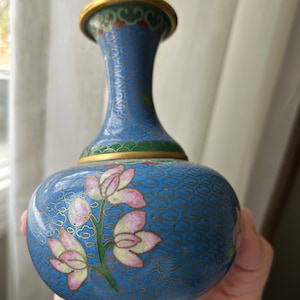 May include: A small blue and green cloisonné vase with a floral design. The vase has a narrow neck and a round body. The floral design features pink and white flowers with green leaves.