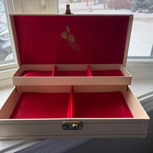 May include: Open, cream-colored jewelry box with a red velvet interior. The box has two levels with compartments and a gold-colored floral design on the inside lid. A gold-colored clasp secures the box.