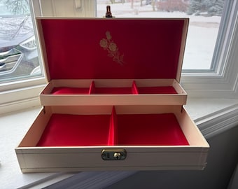 May include: Open, cream-colored jewelry box with a red velvet interior. The box has two levels with compartments and a gold-colored floral design on the inside lid. A gold-colored clasp secures the box.