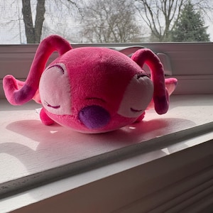 May include: A plush toy in the shape of a pink alien with large ears and a purple patch. The toy is resting on a white windowsill, with a blurred outdoor scene in the background.