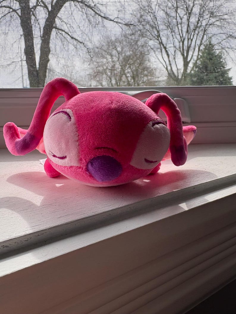 May include: A plush toy in the shape of a pink alien with large ears and a purple patch. The toy is resting on a white windowsill, with a blurred outdoor scene in the background.