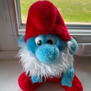 May include: A plush Smurf character toy featuring bright blue skin, a large white beard made of synthetic fur, and a soft red hat. The figure has prominent round plastic eyes with black pupils and is dressed in red trousers that match the hat.