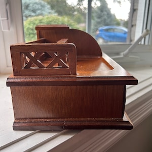 May include: A small, wooden desk organizer with a dark brown finish. The organizer features a sloped back, a small shelf, and a decorative railing. The wood grain is visible, and the piece appears to be well-crafted.