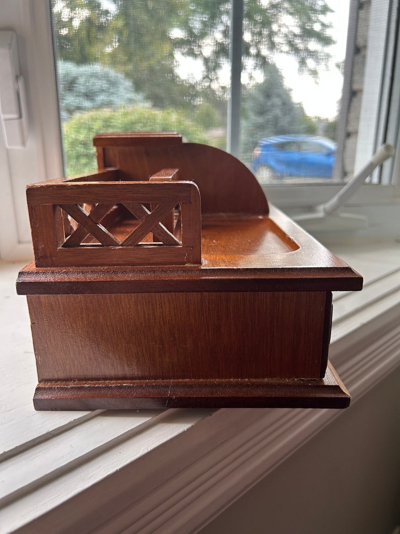 May include: A small, wooden desk organizer with a dark brown finish. The organizer features a sloped back, a small shelf, and a decorative railing. The wood grain is visible, and the piece appears to be well-crafted.