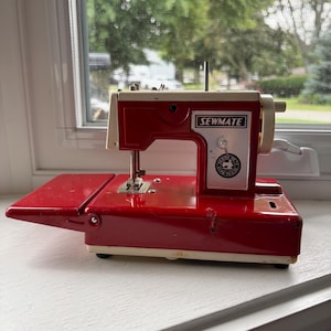 Peut inclure: Une machine à coudre vintage Sewmate avec un corps en métal rouge brillant et crème. L'appareil comprend une table d'extension rabattable, un panneau avant argenté avec le logo Sewmate et un emblème circulaire avec une icône de machine à coudre.