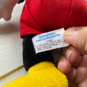 May include: Black and yellow plush Mickey Mouse toy wearing red shorts with a white tag that reads "Disneyland. Walt Disney World" and includes details about the product's materials and origin.