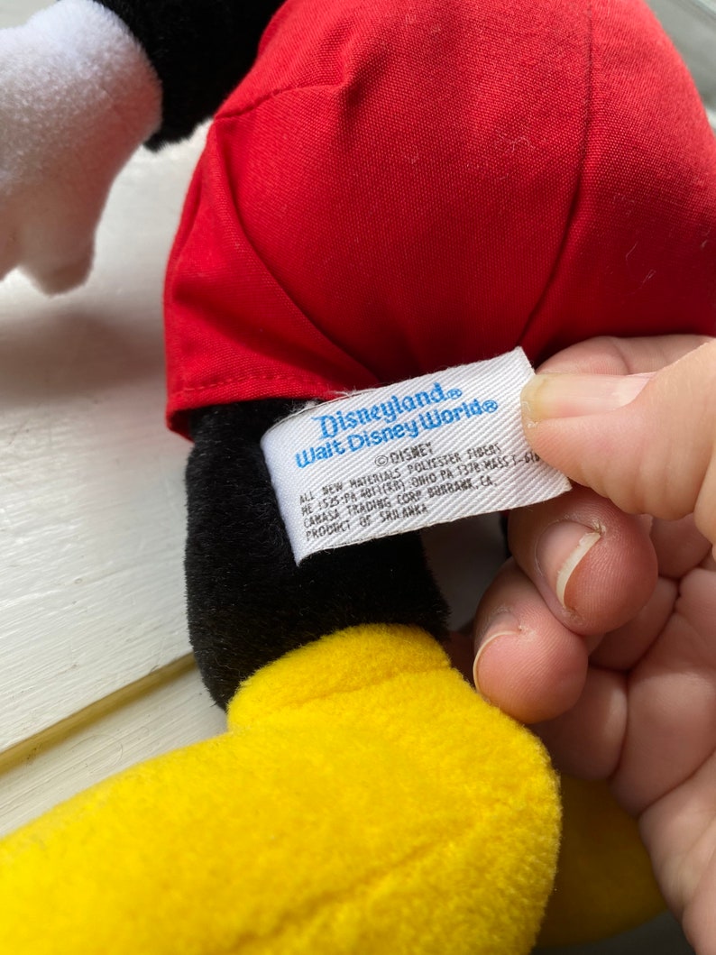 May include: Black and yellow plush Mickey Mouse toy wearing red shorts with a white tag that reads "Disneyland. Walt Disney World" and includes details about the product's materials and origin.