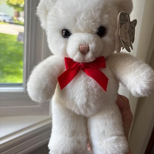 May include: A white teddy bear with a red bow. The bear has black eyes and a light brown nose. A tag is attached to the bear's ear. The bear is a classic soft toy.