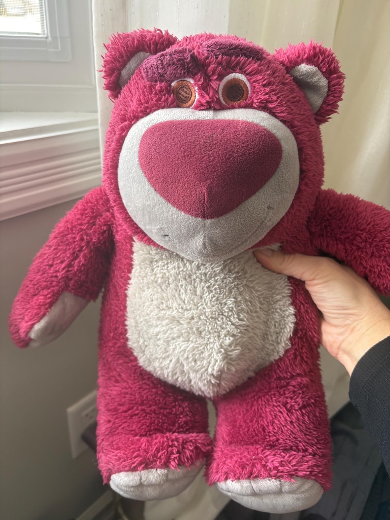 May include: A plush, pink teddy bear with a heart-shaped nose and a white belly. The bear has brown eyes and gray accents on its ears and feet. The toy is soft and fluffy, and appears to be a character from a popular animated film.