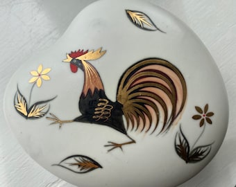 May include: A white ceramic heart-shaped trinket box with a gold and black rooster design. The rooster has a red comb and wattle, and its tail feathers are gold and pink. The box is decorated with gold leaves and flowers.