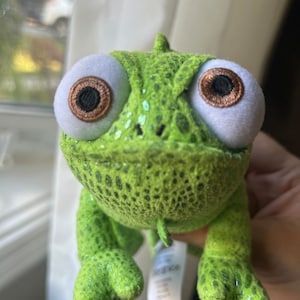 May include: A plush chameleon toy featuring a bright green textured body with a subtle metallic sheen. The creature has two large, round white eyes with brown irises and black pupils, a small snout, and a white fabric tag hanging from its underside.