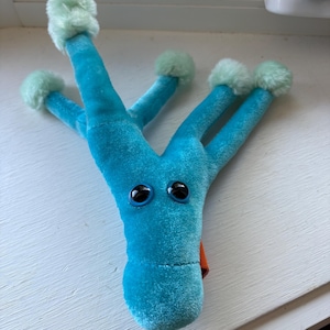 May include: A bright blue plush toy with a Y-shaped body and four long arms. Each arm ends in a soft, light green pom-pom. The toy features two large, round, black plastic eyes and a small orange fabric detail visible on the side of the head.