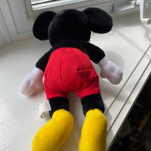 May include: A black and red plush Mickey Mouse toy with yellow shoes. The toy is lying on its back with its arms outstretched.