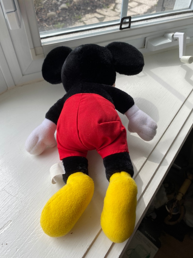 May include: A black and red plush Mickey Mouse toy with yellow shoes. The toy is lying on its back with its arms outstretched.