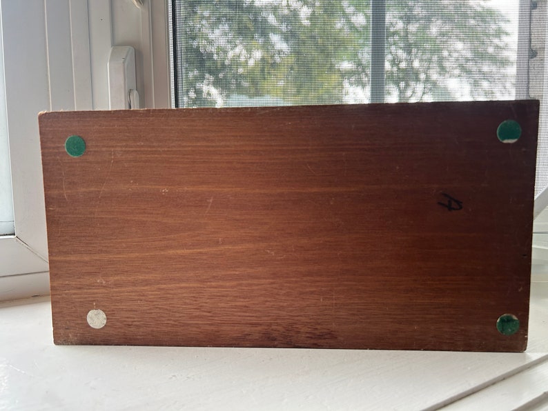May include: A rectangular wooden object with a dark brown finish. The object has four small, circular pads on the corners. The pads are green and white. The object is sitting on a white surface.