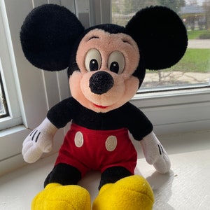 May include: A plush Mickey Mouse toy with black ears, a black body, red shorts with two white buttons, and yellow shoes.