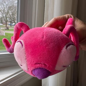 May include: A plush toy in the shape of a pink alien with large, curved antennae. The toy has a rounded head with light pink cheeks, closed eyes, and a purple nose. The toy is held up in front of a window.