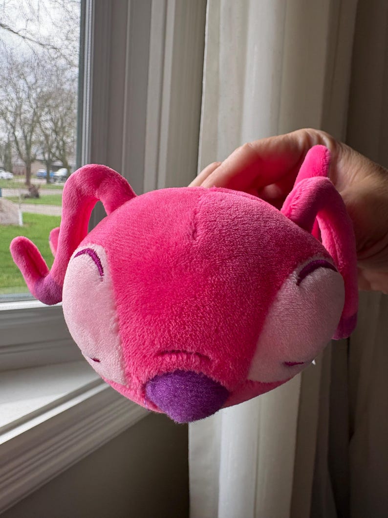 May include: A plush toy in the shape of a pink alien with large, curved antennae. The toy has a rounded head with light pink cheeks, closed eyes, and a purple nose. The toy is held up in front of a window.