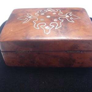 Peut inclure: Une boîte carrée en bois avec un couvercle à charnière. La boîte est de couleur marron chaud avec un motif décoratif incrusté sur le dessus. Le motif est un motif floral blanc stylisé avec un cercle central.