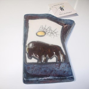 May include: Hand-crafted ceramic art piece featuring a dark brown bison image on a white background, framed by a blue and brown border. A yellow sun and cracked lines are above the bison. The tag reads "Hand Crafted" and "Gary McKague".