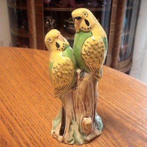 May include: A ceramic ornament depicting two budgerigars perched on a tree stump. The birds have yellow heads and wings, green bodies, and detailed feather patterns. The stump is brown with green and blue accents.