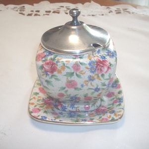 Peut inclure: Un sucrier vintage à motifs floraux avec un couvercle argenté et une soucoupe carrée assortie. Le bol et la soucoupe sont décorés de roses roses et rouges, de fleurs bleues et de feuilles vertes sur fond blanc.