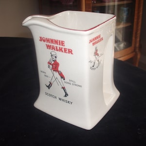 May include: White ceramic jug with a red and black Johnnie Walker logo and a cartoon image of a man walking. The text on the jug reads "Born 1820 - Still Going Strong" and "Scotch Whisky".