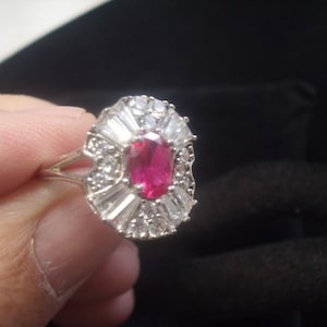 May include: A silver ring with a central oval ruby, encircled by a halo of baguette and round-cut diamonds. The ring's design is symmetrical, with the gemstones arranged in a floral or starburst pattern.