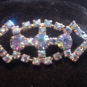 May include: Vintage brooch with an iridescent, multi-coloured design. The brooch features a silver-toned metal setting with a geometric shape, adorned with numerous small, square-cut crystals and larger, round crystals.