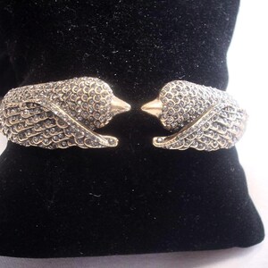 May include: A gold-coloured cuff bracelet featuring two bird heads adorned with crystal-like embellishments. The wings are detailed with smaller crystals. The bracelet is displayed on a black velvet surface.