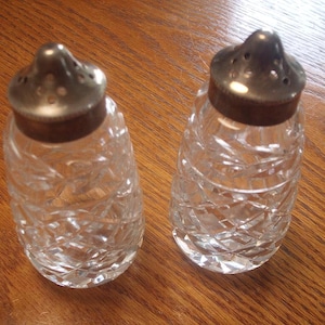 May include: A pair of clear glass salt and pepper shakers with silver-coloured metal lids. The shakers feature a diamond-cut design and are placed on a wooden surface. The lids have small holes for dispensing.