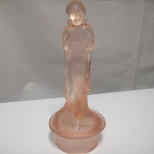 May include: A pink glass figurine of a woman in a long dress, standing on a round base. The figurine has a delicate, frosted appearance and is likely vintage or antique. The figure's arms are crossed in front of her.
