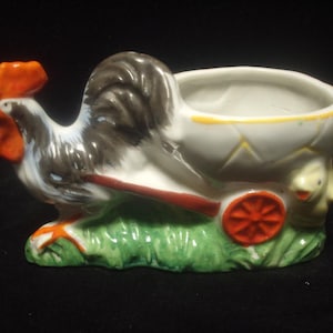 May include: A white ceramic rooster pulling a small white wagon with a red wheel. The rooster has a black and white tail and a red comb. The wagon has a green base and a yellow rim.