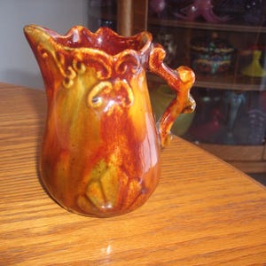 May include: A decorative ceramic jug with a handle, in shades of orange, brown, and yellow. The jug has a ruffled rim and ornate detailing. It is suitable for display or serving, and is a classic piece.