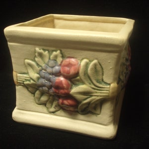 May include: A square ceramic planter featuring a raised design of grapes, leaves, and flowers in shades of green, purple, and pink.