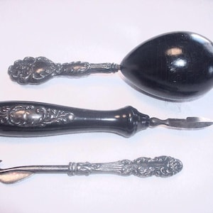 May include: Three antique kitchen tools: an egg separator with a black handle, a can opener, and a knife. The handles are black with silver decorative accents. The egg separator has a large, egg-shaped black head. The can opener and knife have silver blades.