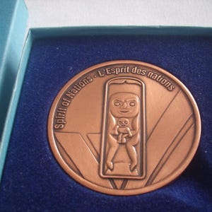 May include: A bronze-coloured commemorative coin with the text "Spirit of Nations - L'Esprit des nations" around the edge. The coin features a stylized figure holding a smaller figure within a rectangular frame. The coin is in a blue velvet-lined box.