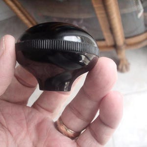 May include: A shiny black, spherical knob with a grooved band around the centre. The knob has a flared base and is held in a person's hand. The background is blurred, showing a hint of furniture.