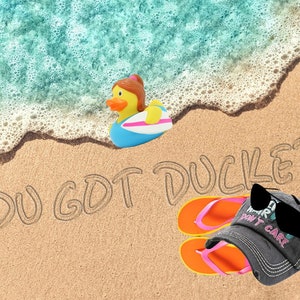 Puede incluir: Una escena de playa con un pato de goma amarillo en una tabla de surf, un cubo azul, una pelota de playa, chanclas, una gorra de b&eacute;isbol negra con el texto "Don't Care" en blanco y las palabras "You Got Ducked" escritas en la arena.