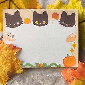 May include: A rectangular notepad with a cream-coloured centre, featuring three black cat faces at the top. The notepad is decorated with autumn-themed illustrations, including candy corn, a caramel apple, a pumpkin, and a cinnamon roll. The notepad is surrounded by yellow leaves.