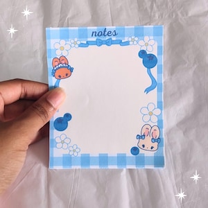 May include: Blue and white checkered notepad with a white border. The border is decorated with white flowers, blue berries, and cartoon bunnies. The notepad has the word "notes" printed at the top.
