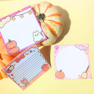 May include: Three square memo pads with Halloween themes. One features a ghost and pumpkins, another has lined paper with a fox and apples, and the third is a circular notepad with a bat, broom, and candy corn. A large pumpkin sits behind them.
