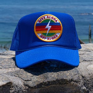 May include: A blue trucker hat with a mesh back and a curved brim. The hat features a circular patch with the words "GIVE ALOHA GET ALOHA" around a lightning bolt design in a rainbow color scheme.