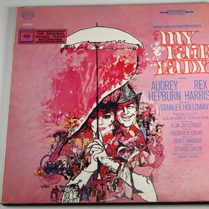 May include: A pink and white vinyl record cover for the musical "My Fair Lady" featuring Audrey Hepburn and Rex Harrison. The cover art depicts a scene from the musical with a large umbrella and a crowd of people.