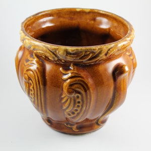 May include: A brown ceramic planter with intricate raised floral designs. The planter has a wide, round opening and a slightly rounded base.