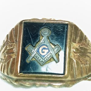 May include: A gold-toned ring with a rectangular black center featuring a Masonic symbol. The symbol includes a compass, square, and the letter "G" in blue. The ring has a detailed, textured design on the sides.