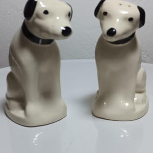 Puede incluir: Un par de saleros y pimenteros de cerámica con forma de perros sentados. Los perros son de color blanco roto con orejas, nariz y collar negros. Cada figura tiene pequeños agujeros en la parte superior de la cabeza para el condimento.